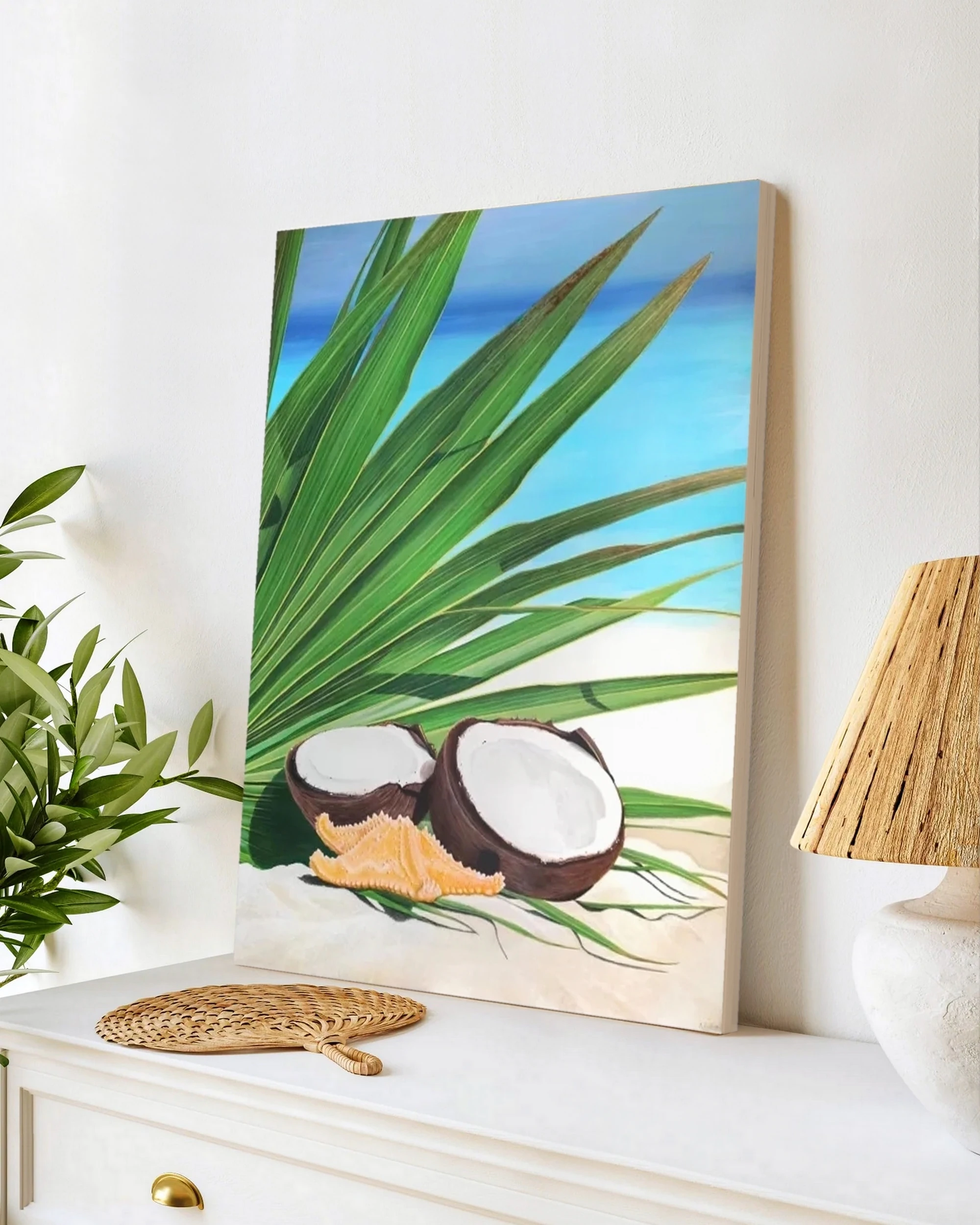 Tableau en peinture d'une noix de coco, étoile de mer et feuille de palmiers sur la plage posée sur un meuble pour une décoration bord de mer tropical exotique