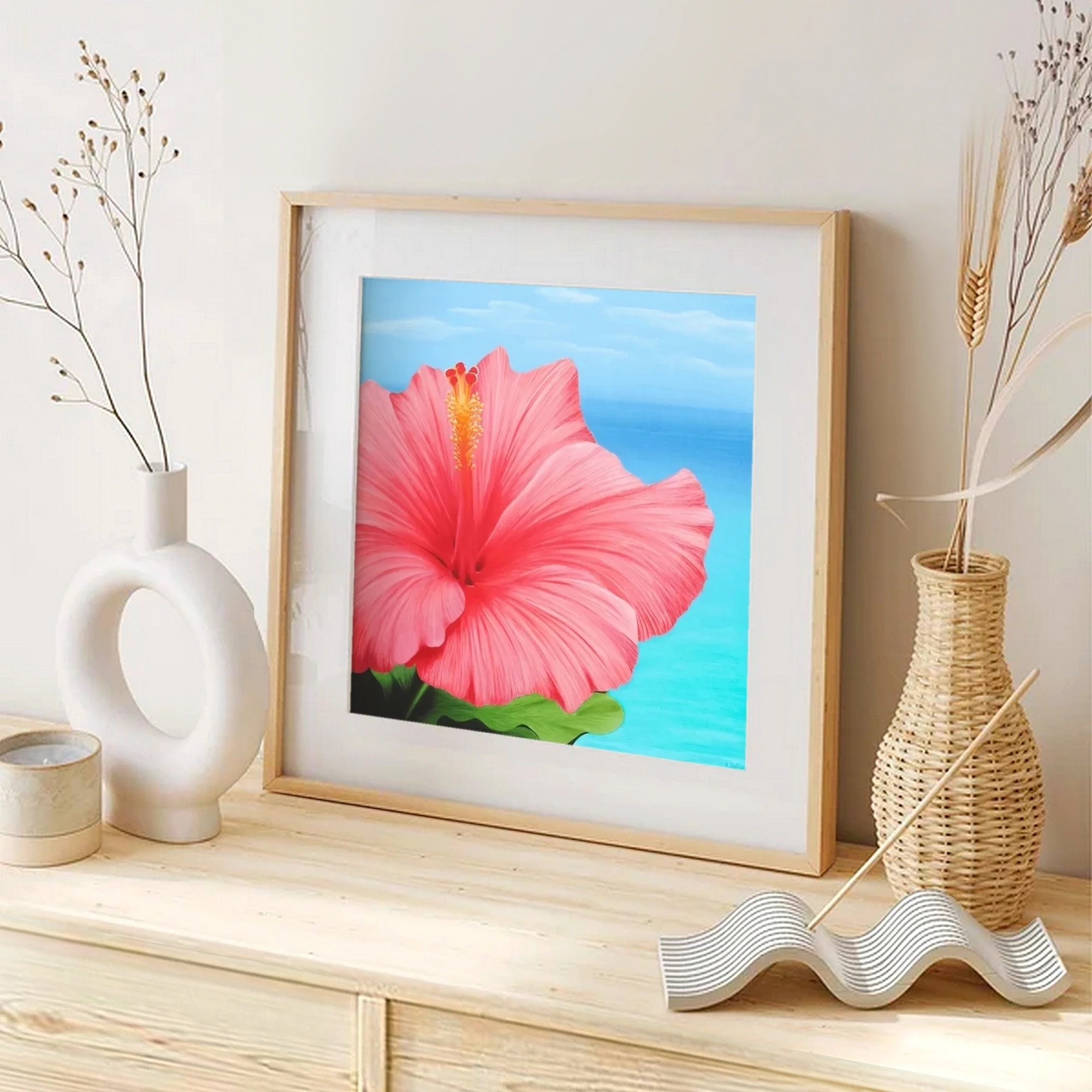 Impression d'art d'une peinture de fleur d'hibiscus rose devant l'océan posée sur un meuble pour une décoration bord de mer tropical exotique