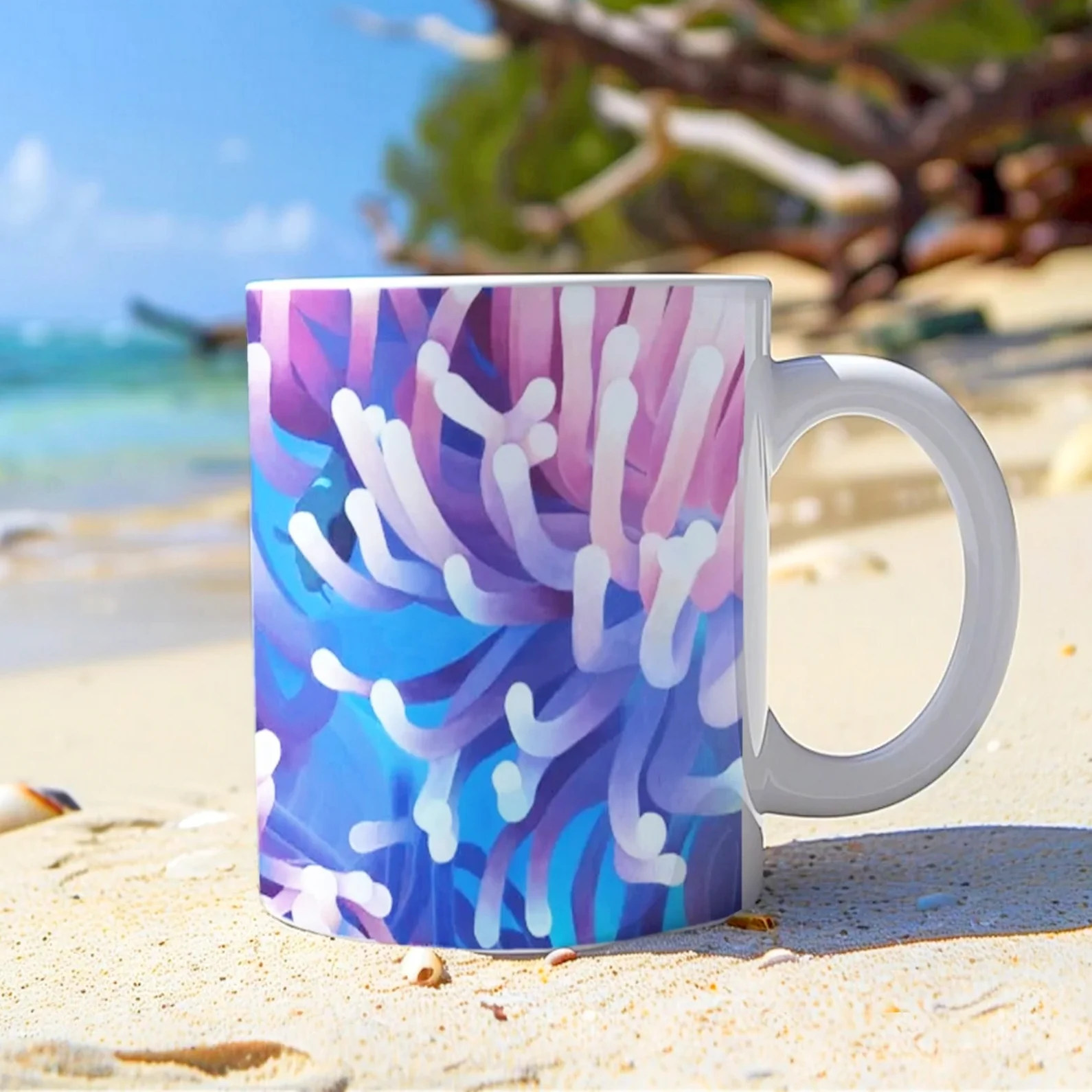 Mug artistique avec anémones de mer et poisson, posé sur le sable avec l’océan en arrière-plan, parfait pour les moments de détente.