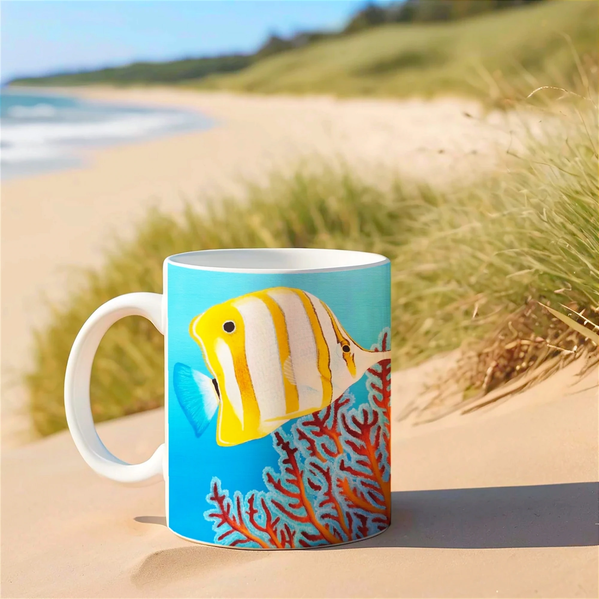 Mug avec poisson Chelmon et récifs marins, posé sur le sable, ajoutant une touche de nature tropicale à votre quotidien.