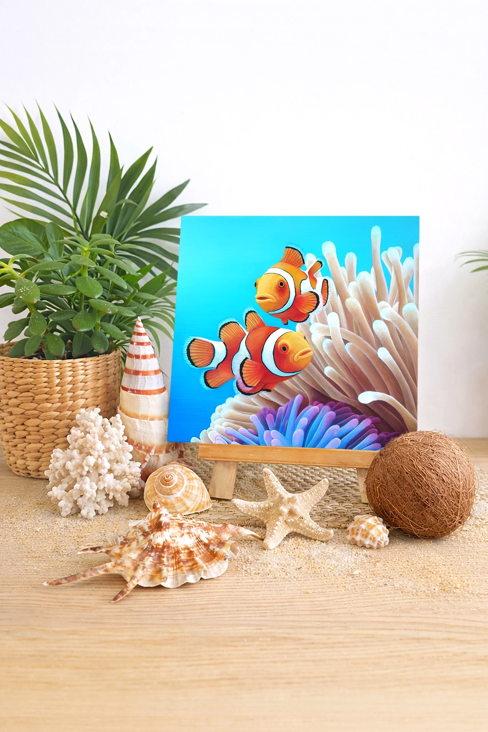 Peinture de deux poissons-clowns dans une anémone posée sur un chevalet entouré de coquillages et de plantes dans une décoration bord de mer.